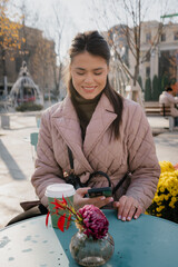Smiling woman in a pink quilted coat sits at an outdoor cafe, checking her phone beside a coffee cup and a vase of flowers.
