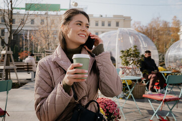 A joyful woman in a padded jacket chats on her cellphone while holding a coffee cup at a sunny outdoor cafe.