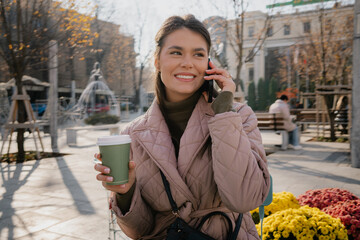 A woman in a pink quilted coat chats on the phone while holding a coffee outdoors.