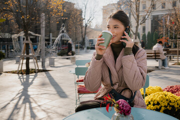 Casual outdoor moment: a woman sips coffee, talks on her phone, with autumn colors and flowers nearby.