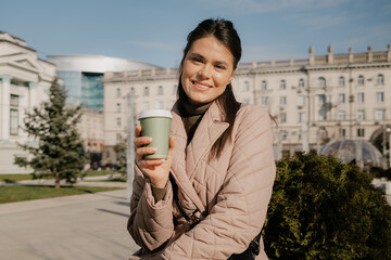 A cheerful woman in a pink quilted jacket enjoys coffee outdoors near urban buildings.