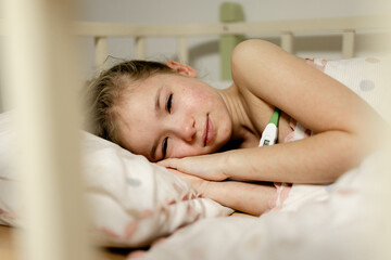 A young girl lying in bed, looking unwell. She has a thermometer in her hand and appears to be resting. The bedding is light-colored with polka dots, and the room has a soft, calm atmosphere.