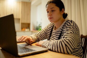 A young woman with a cell phone in hand and a laptop sits at the dining room table in her kitchen in the night light, working online