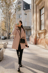 A stylish woman in a pink quilted coat walks along a sunlit city street, sipping coffee.