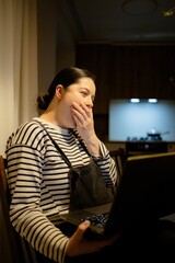 A woman sitting at a wooden table, yawning while using a laptop in a dimly lit kitchen. She wears a striped shirt and has her hair tied back. A vase with flowers is on the table.