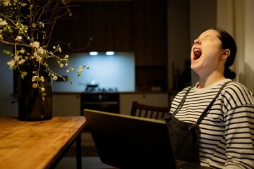 A woman sitting at a table with a laptop, yawning. She is wearing a striped shirt and has her hair tied back. The background shows a kitchen area with soft lighting.