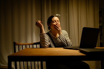 A woman sitting at a wooden table, yawning while using a laptop in a dimly lit kitchen. She wears a striped shirt and has her hair tied back. A vase with flowers is on the table.