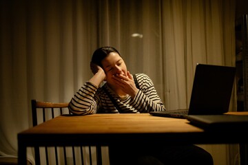 A woman sitting at a table with a laptop, yawning. She is wearing a striped shirt and has her hair tied back. The background shows a kitchen area with soft lighting.
