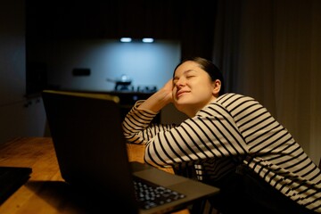 A woman sitting at a table with a laptop. She is wearing a striped shirt and has her hair tied back. The background shows a kitchen area with soft lighting.