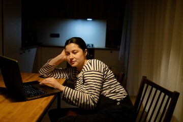 A woman sitting at a table with a laptop. She is wearing a striped shirt and has her hair tied back. The background shows a kitchen area with soft lighting.