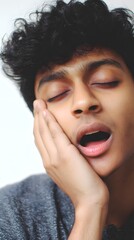 Tired young man with curly hair yawning with his hand on his cheek.