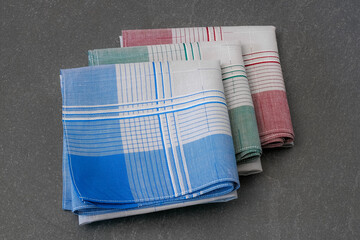 Set of three neatly folded handkerchiefs in various colors (blue, green, and red) with grey centers and a plaid pattern, photographed on a solid gray surface.