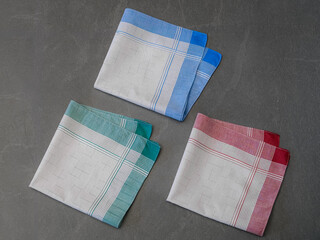 Set of three neatly folded handkerchiefs in various colors (blue, green, and red) with grey centers and a plaid pattern, photographed on a solid gray surface.