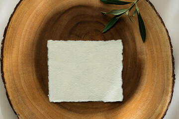 Blank textured paper card on wooden slice with olive branch on beige background. Minimalist mockup with natural light and shadows.	