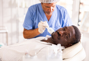 Male cosmetologist performs skin care and maintenance procedure for African man client. Specialist...