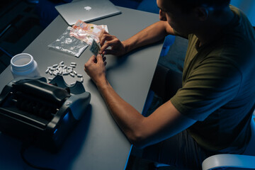 Top view of unrecognizable drug dealer counting Russian rubles preparing drugs, pills, powder narcotics sitting at table with money counting machine in dark room. Concept of drug dealing or addiction.