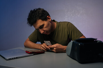 Focused cashier counting banknotes using automatic banknote counter machine at modern dark workplace, sitting at desk with laptop in room illuminated by soft blue light in background.