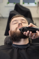 Woman Barber wielding electric hair clipper shaving beard customer in modern barbershop. Bearded Man client visit gentleman grooming beauty salon for hairstyle care. Male beard style concept