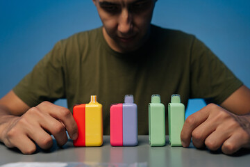 Blurred male holding and presenting various new heat-not-burn tobacco heating systems in vibrant colors on gray table, against isolated blue background.