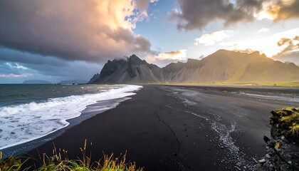 Dramatic sunset over a black sand beach, framed by dramatic mountains and stormy clouds.