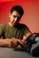 Vertical studio portrait of serious focused cashier counting stack of banknotes using professional electronic money counting machine with red light on red isolated background.