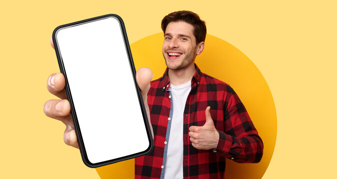 A cheerful man wearing a red and black checkered shirt shows a blank smartphone screen. He stands in front of a bright yellow backdrop, giving a thumbs up.