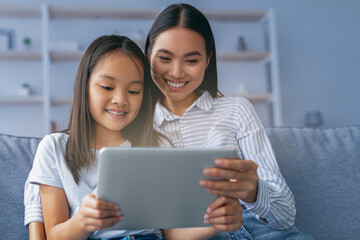 Happy asian mother and child girl using digital tablet together while sitting on couch at home, free space. Loving mom and daughter playing online games on pad or surfing internet