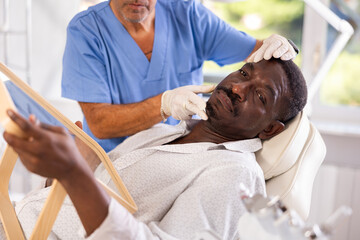 Experienced male cosmetologist examines facial skin of African-American man patient and advises on...