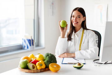 Obraz premium Portrait of happy woman dietitian holding apple, sitting at desk in clinic office with plate of fruits and vegetables, recommending healthy eating