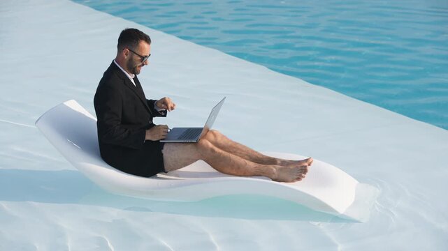 Businessman in glasses, swim trunks, and blazer holding a video call while lying on a poolside lounger. Concept remote communication and modern work flexibility.