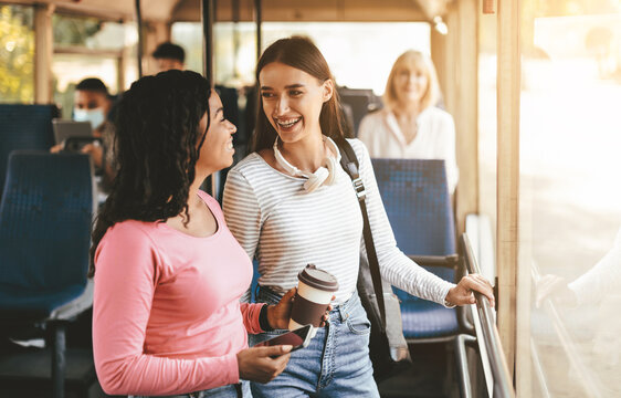 Two friends engage in a lively conversation on a city bus in the afternoon. One of them holds a coffee cup while both are smiling and sharing a light moment.
