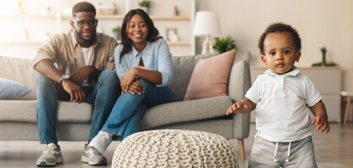 Childhood And Parenthood Concept. Cute baby toddler standing up in living room making first steps, cheerful African American parents sitting on the sofa looking at son trying to walk