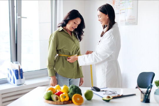Professional woman dietologist finding out waist length of mature female patient with measuring tape in consulting room in clinic