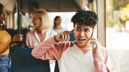 A young man sits on a bus talking on his phone, showing concern on his face. Other passengers occupy the bus, suggesting a busy afternoon commute.