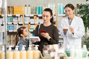 Woman in thirties researching ingredients of vitamins online via smartphone before buying for preteen daughter at pharmacy while friendly professional female pharmacist assisting in background..