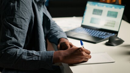 Remote job professional at his personal office balancing activities like research, writing notes and reading articles. Young freelancer uses laptop for project planning and data gathering. Camera B.