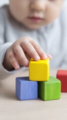 Baby hand places bright yellow block on blue green wooden blocks, building tower