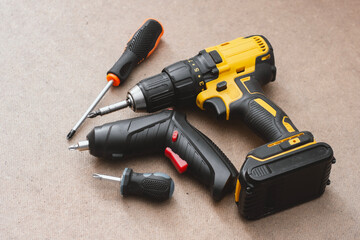 Set power tools including yellow cordless drill with battery pack, electric screwdriver, and manual screwdrivers handles on a wooden background. DIY and construction equipment.