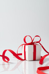 A small square white gift box tied with a red satin ribbon that forms a perfect bow on top, for Christmas, New Year or birthday theme. The ribbon trails around the box on a reflective white surface
