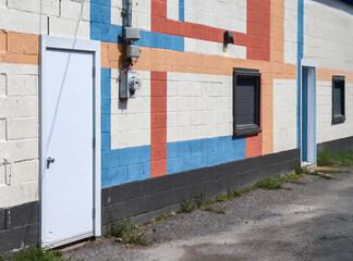 Building exterior facade, colourful painted cinder block wall, white metal exterior doors, two boarded up windows, electric meter, daytime, nobody