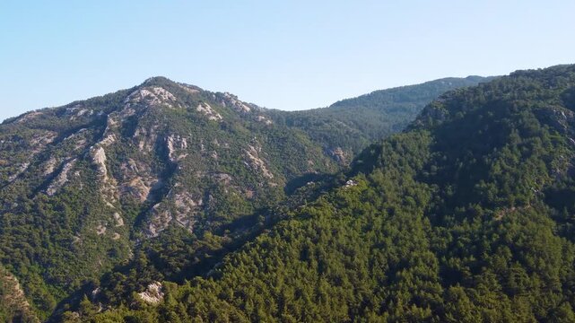 An aerial view showcases the vibrant, lush green mountains and dense forests surrounding Turunc, a picturesque coastal town in Turkey. The clear blue sky provides a serene backdrop to the rugged, verd