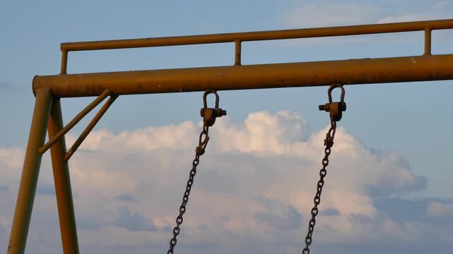 Close-up of an old swing set structure with rusty chains hanging under a cloudy sky. A nostalgic, symbolic view of childhood memories, time passing, and the contrast between play and decay.