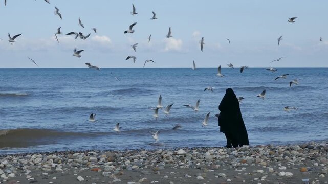 A Muslim woman in black chador stands on the rocky shore, feeding seagulls as they circle above the waves. A poetic and cinematic seascape blending culture, faith, and nature&rsquo;s freedom.