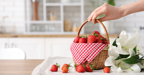 Female hand holding wicker basket with tasty strawberries and lily flowers on table in kitchen