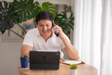 Asian freelancer talking on phone and taking notes while working on tablet at home