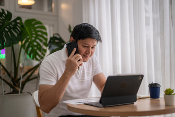 Asian freelancer talking on phone and working on tablet in cozy cafe