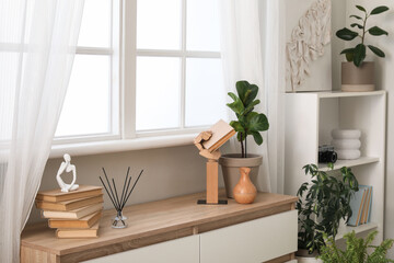 Books and statuette on chest of drawers near window in stylish room, closeup