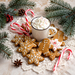 “White holiday mug of hot chocolate with whipped cream, gingerbread cookies, candy canes, and festive pine branches on a warm textured Christmas background”
