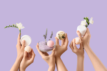 Female hands holding bath bombs, flowers, bowl with sea salt and jars of cosmetic products on lilac...