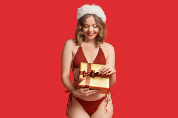 Attractive young woman in swimsuit with Santa hat and Christmas gift on red background